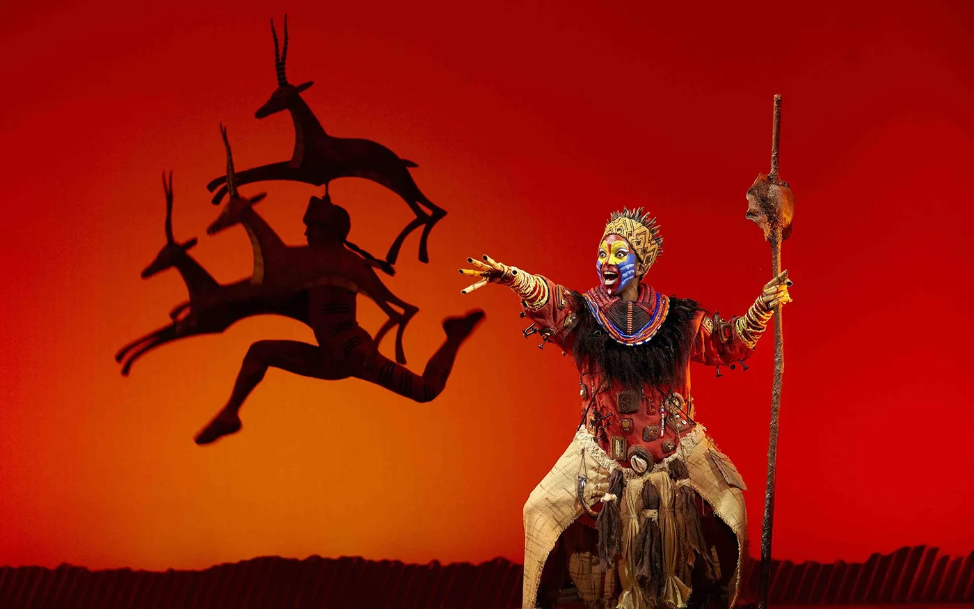 A performer holding a staff against a red El Rey León stage backdrop