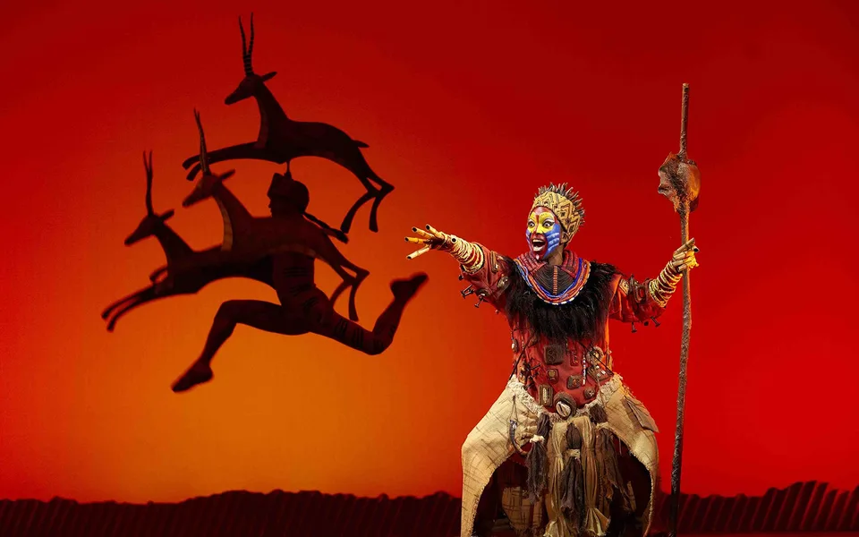 A performer holding a staff against a red El Rey León stage backdrop